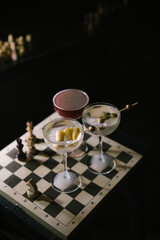 Three Martini cocktails, two differently garnished dirty martinis and a twist on espresso martini, standing on a wooden chess board in the dark room, close up view 