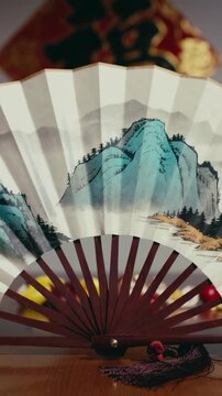 Vertical video: Opening fan pivoting at rivet, revealing blue-green mountain on table with tassel