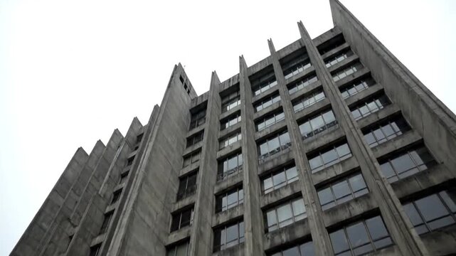 A dramatic low-angle shot captures the imposing facade of a gray concrete Brutalist high-rise building against a stark white sky, emphasizing its repetitive windows and severe vertical lines over the