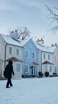 Vertical video: Moving man in dark coat entering left, passing pastel houses on street, copy space