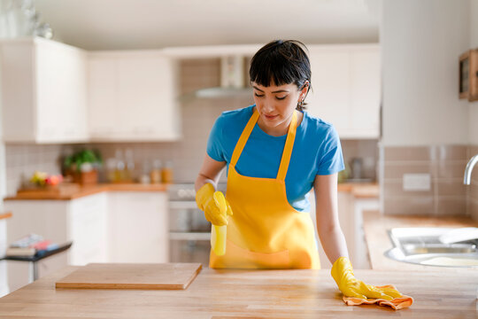Woman cleaning kitchen counter with spray bottle and cloth in modern home during afternoon