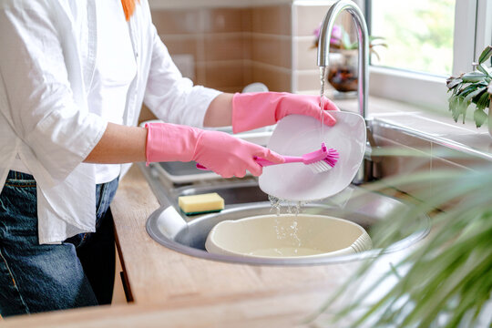 Woman washes dishes in a kitchen at home using a brush and wearing gloves during the day