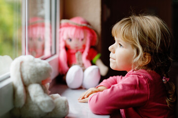 Cute toddler girl sitting by window and looking out on rainy day. Dreaming child with doll and soft...