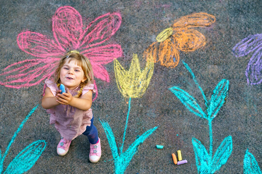 Toddler girl drawing colorful flowers with sidewalk chalk outdoors, top view of creative play on pavement showing carefree childhood, real people