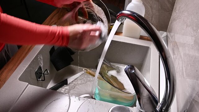 A close-up with localized lighting. The hands of a Caucasian woman washing dishes in a home kitchen late at night in the dark.