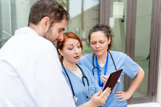 Medical team collaborating on tablet, discussing healthcare data and patient information outdoors near a hospital
