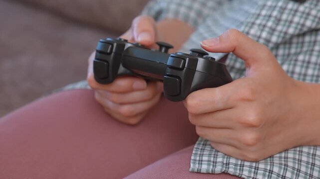 Unrecognizable woman holding a joystick and playing video games on a console at home, pressing the buttons on the controller and having fun during her leisure time on the couch