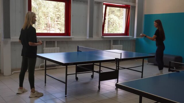 Two active young women enthusiastically playing a competitive game of table tennis indoors, hitting the ball with red rackets during a friendly match in a sports club room
