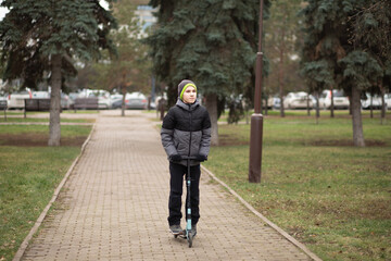 Fototapeta premium A child in a gray hooded jacket and black pants rides a blue kick scooter on a paved park path surrounded by trees and green grass.