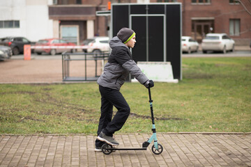 A child in a gray hooded jacket and black pants rides a blue kick scooter on a paved park path surrounded by trees and green grass.