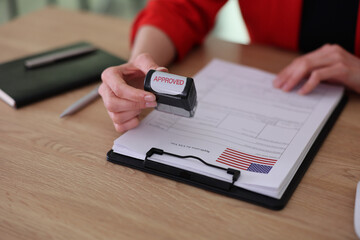 A person holds a rubber stamp with the word approved, poised to complete important paperwork. Sunlight streams through the window, creating a warm atmosphere in the office.