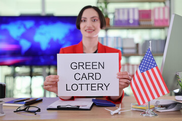 A confident woman in a vibrant red blazer holds a sign announcing the green card lottery. Behind her, shelves filled with documents and an American flag symbolize dreams of new beginnings in the U.S.