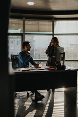 Two business people work together in a bright office, chatting beside laptops and documents. A man and a woman converse at the desk as coffee breaks and everyday teamwork unfold.