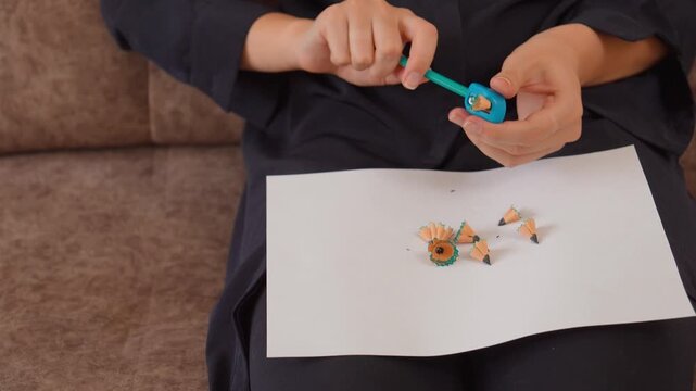 Close-up of female hands sharpening a graphite pencil with a blue sharpener, with shavings and broken tips falling onto a white paper sheet, representing preparation for drawing or studying