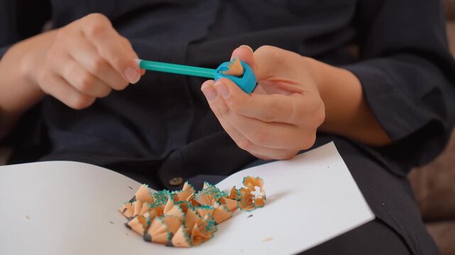Close up view of a person's hands holding a pencil sharpener and carefully sharpening a turquoise colored pencil, with the wood and graphite shavings collecting on a piece of white paper