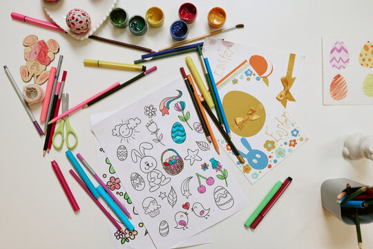 Children creating Easter-themed drawings and coloring pages with colored pencils, markers, and paints on table, hands of Caucasian kids visible while working on festive crafts