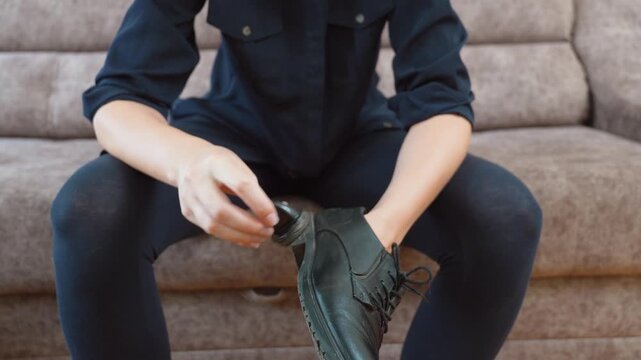 Female hands applying polish and cleaning dirty black leather shoes with a special sponge brush while sitting on a couch at home, a domestic chore of footwear care and maintenance