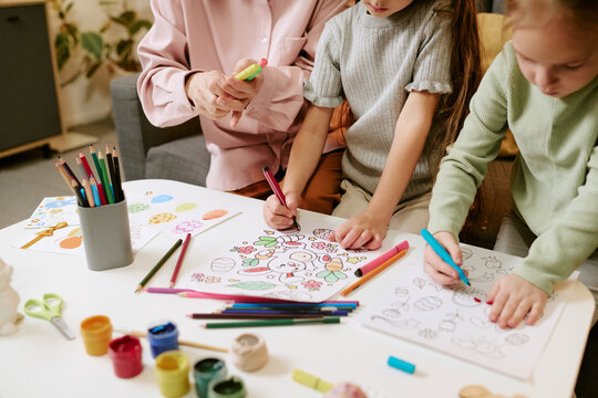 Two Caucasian girls drawing and coloring Easter-themed pictures with colored pencils and markers while senior Caucasian woman assisting, art supplies scattered on table