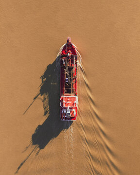 Aerial view of a red barge casting dark shadows on the muddy waters, a stark contrast of color and form in motion, Cite des Pains, Nouvelle-Aquitaine, France.