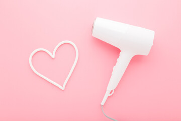 Heart shape and new white female hair dryer on light pink table background. Pastel color. Closeup. Care about hair. Top down view.