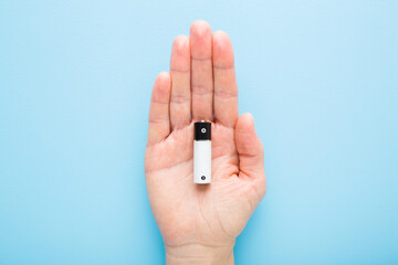 Young adult woman palm holding and showing new white black battery on light blue table background. Pastel color. Top down view.
