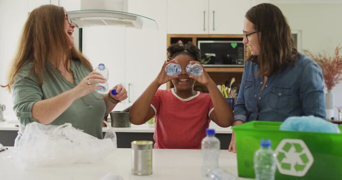 Happy caucasian lesbian couple and their african american daughter sorting waste in kitchen