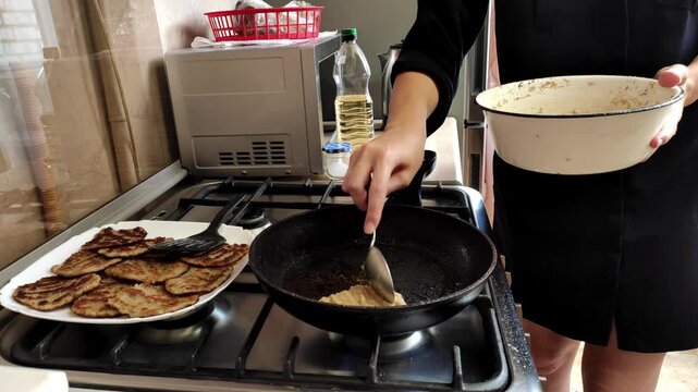 Close-up view of a woman's hands using a spatula to fry traditional golden potato pancakes, or draniki, in hot oil on a gas stove, preparing a delicious homemade meal in the kitchen