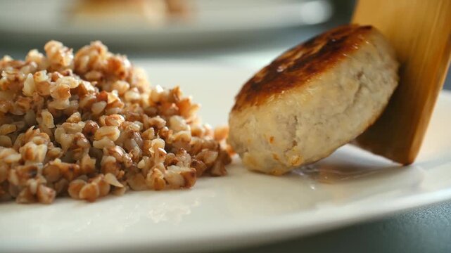 Close up wooden spatula placing golden fried chicken cutlet on white plate next to serving of healthy boiled buckwheat porridge, preparing delicious homemade lunch or dinner. Fried chicken cutlet 4k
