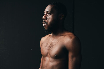 Fototapeta premium African man with dark skin and muscular body posing shirtless in creative conceptual visual style on black background. Portrait with dramatic lighting and serious expression in studio.