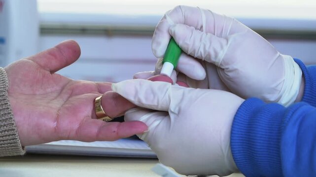 Close up view of a medical professional in white protective gloves pricking a patient's finger with a green lancet device to draw blood for diagnostic testing in a clinical setting