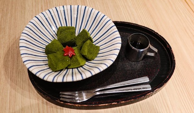 close up of traditional matcha warabi mochi cubes drizzled with kuromitsu black sugar syrup, garnished with red maple leaf decoration served in striped Japanese bowl on lacquer tray, with copyspace