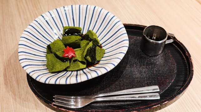 close up of traditional matcha warabi mochi cubes drizzled with kuromitsu black sugar syrup, garnished with red maple leaf decoration served in striped Japanese bowl on lacquer tray, with copyspace