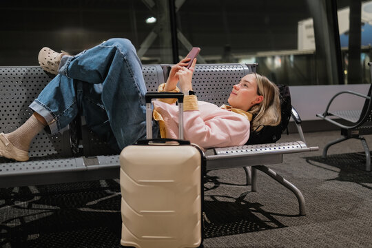 Airport layover, exhausted female traveler sleeping on terminal bench with suitcase and backpack, delayed flight, overnight wait, travel fatigue, jet lag, solo travel concept.