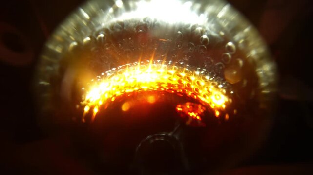 A close-up of a vessel with a bright orange-red element at the base, surrounded by rising bubbles and glittering condensation on the glass surface. The dark background with soft, diffused light source
