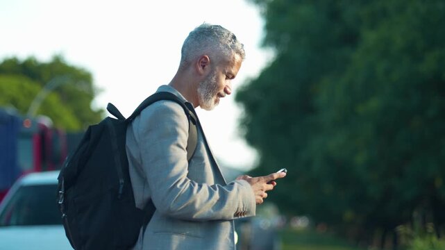Close up of mature Brazilian businessman with backpack using smartphone crossing street with stopped traffic in city