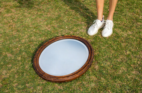 Old vintage oval mirror with wooden frame lying on green grass near female legs in white sneakers, outdoor reflection concept, minimalist surreal scene with copy space.