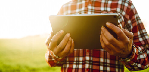 A man stands in a green field at sunset, holding a tablet. The sunlight creates a warm glow. The man is wearing a plaid shirt and appears to be staring intently at the device. © maxbelchenko