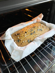 Freshly baked whole grain bread with seeds in a parchment-lined loaf pan inside an oven, showcasing golden crust and warm lighting. Gluten-free home baking concept, green buckwheat bread