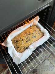Freshly baked whole grain bread with seeds in a parchment-lined loaf pan inside an oven, showcasing golden crust and warm lighting. Gluten-free home baking concept, green buckwheat bread