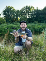 A bearded fisherman in the grass by the river, showing off his catch on the bank of a summer river lined with dense trees. Fishing, wilderness recreation, active recreation