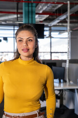 Fototapeta premium Woman standing, glancing right of camera in office with windows and monitors, wearing mustard top
