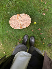 Person wearing black rubber boots stands on grass beside a woven basket filled with mushrooms and surrounded by fallen leaves in a natural outdoor setting