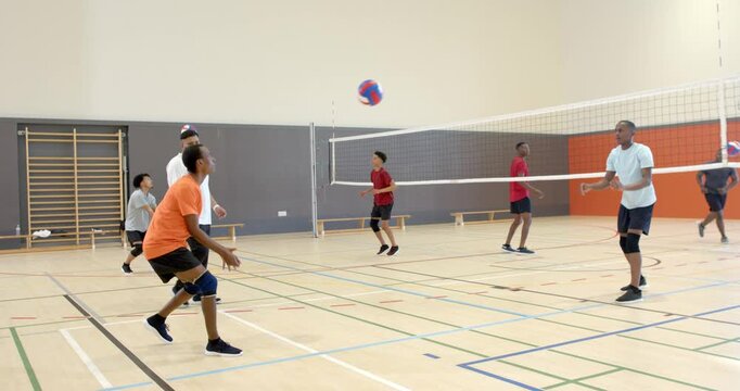 Diverse male team initiating rally by tossing volleyball over net, exchanging bumps at school gym