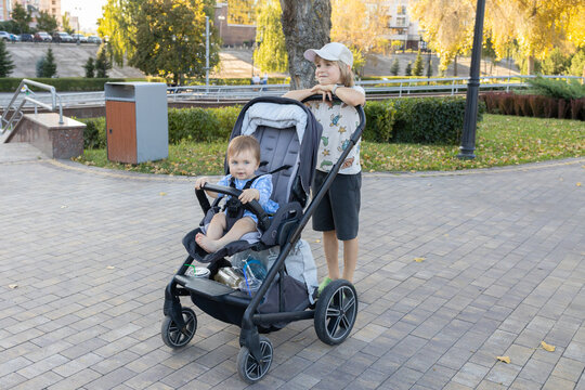 An older brother pushes a baby sitting in a stroller. The siblings are walking along a city street on a summer day, the older one babysitting the younger one.