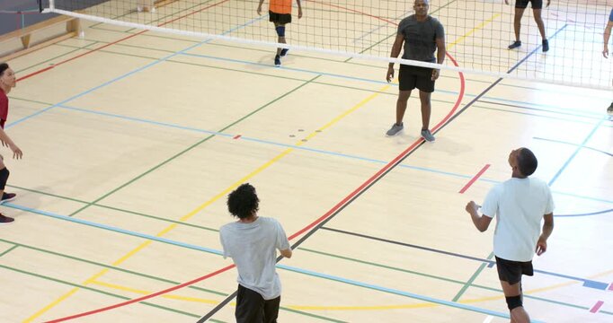 Gray-shirt male server serving ball over net, red-shirt male attacker spiking into blocker at gym