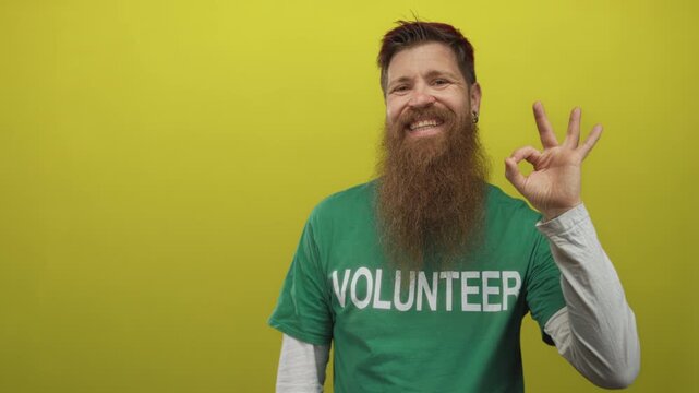 Young man with long beard wearing volunteer shirt makes ok sign in yellow studio; approval generosity.