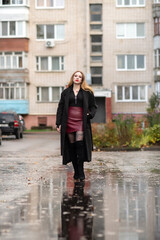 Obraz premium Young woman in black coat and knee-high boots walks through a rain-soaked street with colorful autumn foliage and reflections visible on the wet pavement