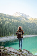 Naklejka premium A female traveler, wearing a hat and carrying a backpack, enjoys nature while sitting on a rock by a lake. A beautiful woman admires the mountain scenery at sunset. Concepts of adventure and freedom.