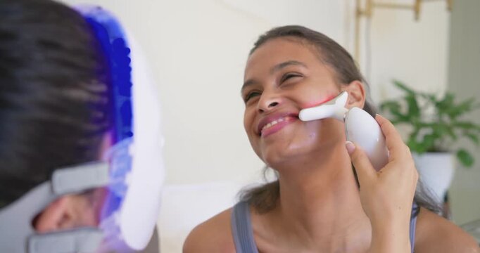 Diverse female operator placing red-light device on client wearing LED mask for skincare at home