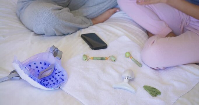 Female friends picking clear serum bottle on bed, passing rollers and applying facial treatment